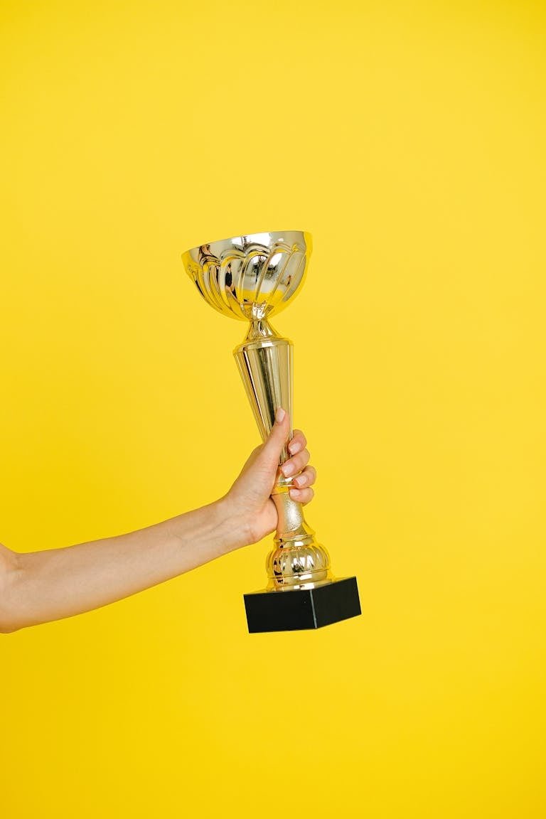 A hand holding a gold trophy against a solid bright yellow background, symbolizing victory.
