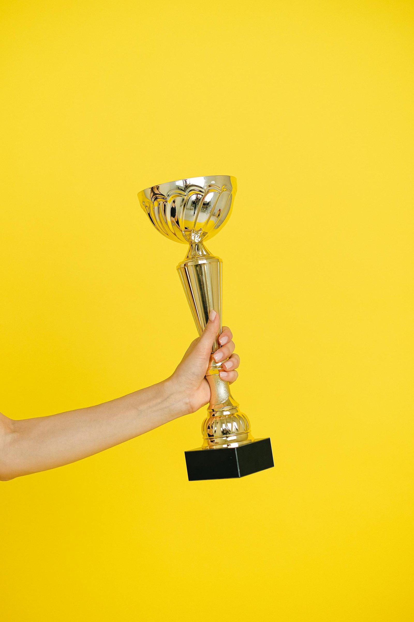 A hand holding a gold trophy against a solid bright yellow background, symbolizing victory.